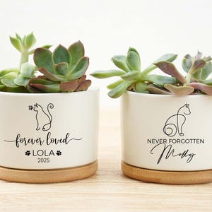May include: Two white ceramic planters with succulents, each on a wooden base. One planter reads "forever loved" with a cat silhouette, the name "LOLA", and the year "2025". The other reads "never forgotten" with a cat silhouette and the name "Molly".