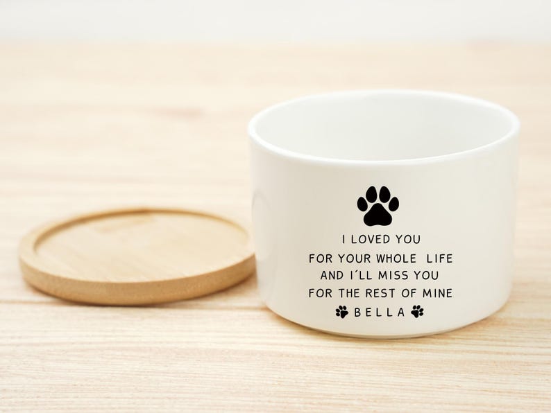 May include: White ceramic pet bowl with a black paw print and the text "I LOVED YOU FOR YOUR WHOLE LIFE AND I'LL MISS YOU FOR THE REST OF MINE BELLA". A wooden coaster is next to the bowl.