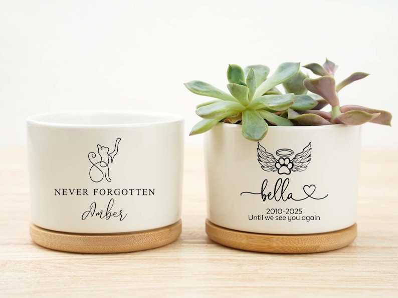 May include: Two white ceramic planters with bamboo bases. One features a cat silhouette and the text "NEVER FORGOTTEN Amber". The other has a paw print with wings and the name "Bella" with dates and the phrase "Until we see you again".