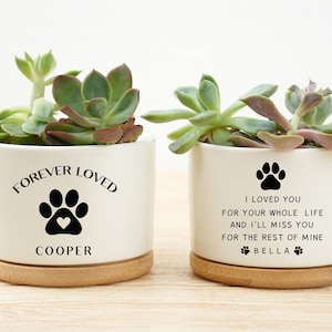 Peut inclure: Deux pots de fleurs en céramique blanche avec des succulentes, chacun sur une base en bois. L'un porte l'inscription "FOREVER LOVED" avec une empreinte de patte et un cœur, et le nom "COOPER". L'autre porte "I LOVED YOU" avec une empreinte de patte et le nom "BELLA".