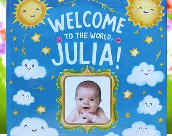 Könnte beinhalten: Ein blaues Babybuch mit einem goldenen Rahmen und einem Foto eines Babys. Das Buch trägt den Text "Willkommen auf der Welt, Julia!" auf der Vorderseite.