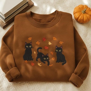 May include: A brown sweatshirt with an embroidered design of three black cats and autumn leaves. The cats have yellow eyes and are in different poses. The sweatshirt has a crew neck and cuffed sleeves. A small orange pumpkin is in the background.