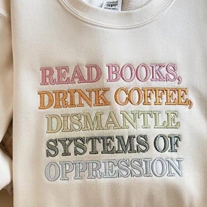 Op de afbeelding: Cr&egrave;mekleurige sweatshirt met geborduurde tekst in diverse kleuren. De tekst luidt: "READ BOOKS, DRINK COFFEE, DISMANTLE SYSTEMS OF OPPRESSION." De sweatshirt heeft lange mouwen en een ronde hals.