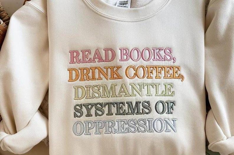 Op de afbeelding: Cr&egrave;mekleurige sweatshirt met geborduurde tekst in diverse kleuren. De tekst luidt: "READ BOOKS, DRINK COFFEE, DISMANTLE SYSTEMS OF OPPRESSION." De sweatshirt heeft lange mouwen en een ronde hals.