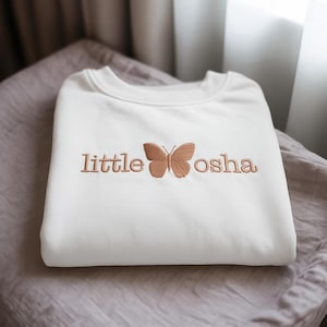 以下が含まれることがあります： 「little osha」の刺繍と茶色の蝶のデザインが施された白いクルーネックのスウェットシャツ。スウェットシャツは、柔らかく質感のある表面にきれいに折りたたまれています。