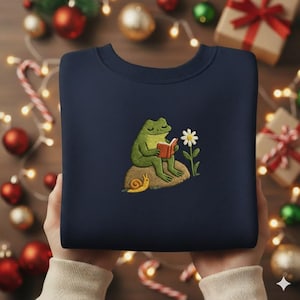 Könnte beinhalten: Marineblaues Sweatshirt mit einer Stickerei einer grünen Froschfigur, die ein Buch liest. Eine weiße Gänseblümchenblume und eine gelbe Schnecke sind ebenfalls auf dem Sweatshirt gestickt. Das Sweatshirt ist gefaltet und wird von einer Person gehalten.