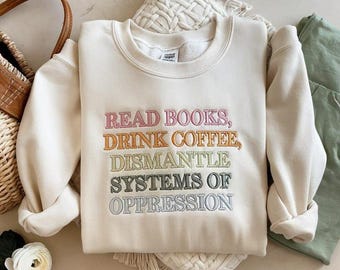 May include: Cream-colored sweatshirt with embroidered text in various colors. The text reads "READ BOOKS, DRINK COFFEE, DISMANTLE SYSTEMS OF OPPRESSION." The sweatshirt is folded, with a woven bag and a white flower in the background.