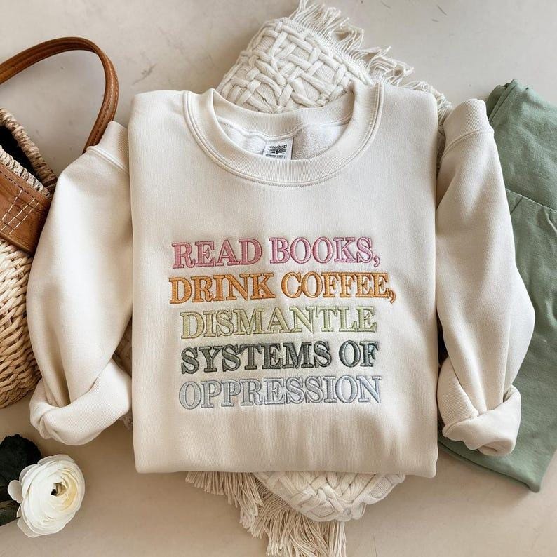 Op de afbeelding: Cr&egrave;mekleurige sweatshirt met veelkleurig geborduurde tekst. De tekst luidt: "READ BOOKS, DRINK COFFEE, DISMANTLE SYSTEMS OF OPPRESSION." De sweatshirt is opgevouwen, met een geweven tas en een witte bloem op de achtergrond.