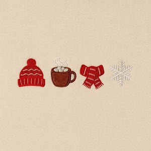May include: Embroidered winter-themed design featuring a red knit cap with a pom-pom, a brown mug of hot chocolate with marshmallows, a red scarf, and a white snowflake. The design is on a cream-colored background.