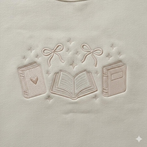 May include: Cream-colored sweatshirt with embroidered design. Features three books, two bows, and star-shaped accents. One book has a heart, and another is open. The embroidery is in a soft pink.