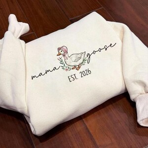 May include: Cream-colored sweatshirt featuring an embroidered goose wearing a pink bonnet and floral accents. The words "mama goose" and "EST. 2026" are also embroidered on the front. The sweatshirt has ribbed cuffs and a relaxed fit.
