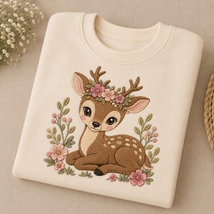 May include: A cream-coloured crewneck sweatshirt featuring a central embroidered design of a brown fawn lying down. The fawn has a floral crown of pink and white flowers on its head and is surrounded by green leaves and small pink flower accents.