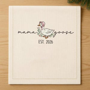 May include: Cream-colored linen napkin with a stitched border. The center features a whimsical illustration of a goose wearing a pink bonnet, surrounded by floral accents. The words "mama goose" and "EST. 2026" are written in script.