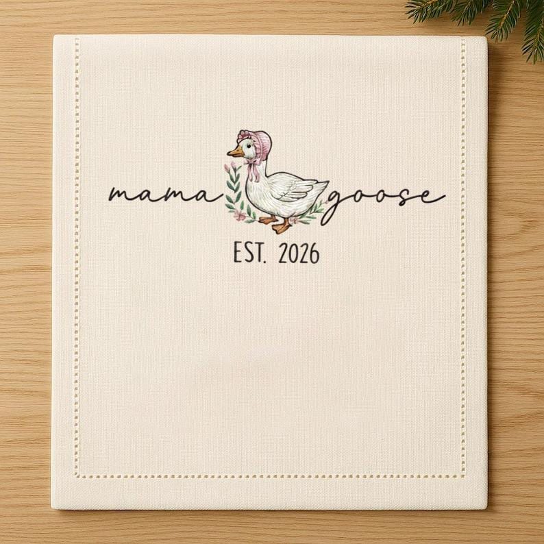 May include: Cream-colored linen napkin with a stitched border. The center features a whimsical illustration of a goose wearing a pink bonnet, surrounded by floral accents. The words "mama goose" and "EST. 2026" are written in script.