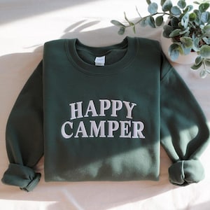May include: A dark green sweatshirt with the words "HAPPY CAMPER" embroidered in white across the front. The sweatshirt has a crew neck and long sleeves with rolled-up cuffs. The garment is folded neatly on a white surface.