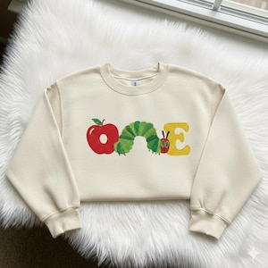 May include: Cream-colored sweatshirt with the word "ONE" in a colorful design. The "O" is a red apple, the "N" is a green caterpillar, and the "E" is yellow. The sweatshirt is made of a soft material.