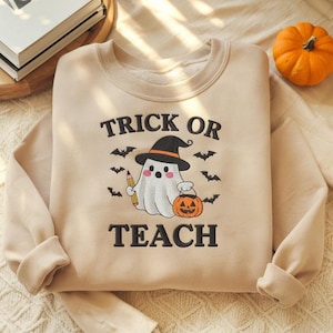 Könnte beinhalten: Beiges Sweatshirt mit dem Text "TRICK OR TEACH" und einer Grafik eines Geistes mit Hexenhut, der einen Bleistift und einen Kürbis hält. Schwarze Fledermäuse sind um den Geist herum verstreut. Im Hintergrund ein kleiner orangefarbener Kürbis.