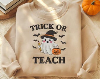 May include: Beige sweatshirt with the text "TRICK OR TEACH" and a graphic of a ghost wearing a witch hat, holding a pencil and a jack-o'-lantern. Black bats are scattered around the ghost. A small orange pumpkin is in the background.