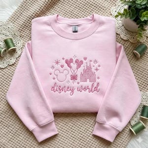May include: A light pink crewneck sweatshirt featuring centered embroidery in a darker pink thread. The design includes a castle, a Mickey Mouse head outline, and balloon shapes, with the text "disney world" written in a cursive script below.