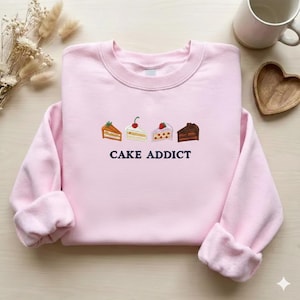 Pode incluir: Um moletom rosa claro com a frase bordada "CAKE ADDICT" abaixo de quatro fatias de bolo bordadas. Os bolos são decorados com cerejas, morangos e glacê.
