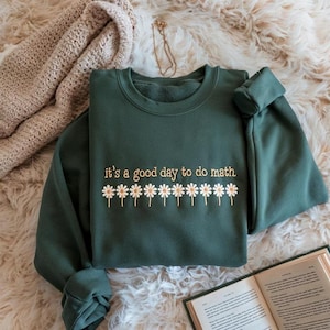 Könnte beinhalten: Ein dunkelgrünes Sweatshirt mit dem gestickten Satz "it's a good day to do math" über einer Reihe von Gänseblümchen. Das Sweatshirt liegt auf einer flauschigen, cremefarbenen Oberfläche.