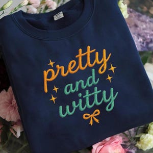 Puede incluir: Una sudadera azul marino con el texto bordado "pretty and witty" en naranja y verde. El diseño incluye estrellas y un pequeño lazo. La sudadera está doblada y rodeada de flores.