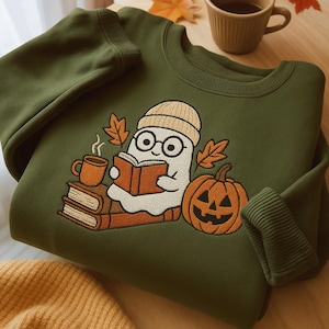 Puede incluir: Sudadera verde oliva con un bordado de un fantasma leyendo un libro, con gorro y gafas. El diseño incluye una calabaza, una taza de café y hojas de otoño. La sudadera está doblada sobre una superficie de madera.