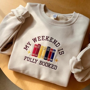 以下が含まれることがあります： クリーム色のスウェットシャツ。刺繍された「MY WEEKEND IS FULLY BOOKED」の文字と、カラフルな本が並んでいます。本は赤、黄色、青です。スウェットシャツは、リブ編みの袖口が付いた長袖です。