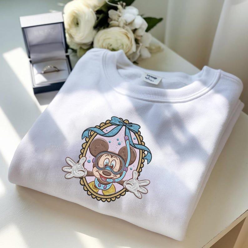 May include: White sweatshirt featuring a colorful embroidered design of Mickey Mouse wearing a snorkel and diving mask. The design is framed in a decorative oval with a blue bow. The sweatshirt is folded neatly.