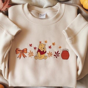 May include: Cream-coloured sweatshirt with an embroidered design of Winnie the Pooh, a bow, a honey pot, and autumn leaves. The design is in shades of orange, red, and yellow. The sweatshirt has a crew neck and long sleeves.