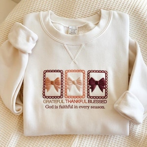 May include: A cream-coloured sweatshirt featuring embroidered bows in three frames. The bows are in shades of brown, pink, and burgundy. The text "Grateful Thankful Blessed" and "God is faithful in every season" are embroidered below.