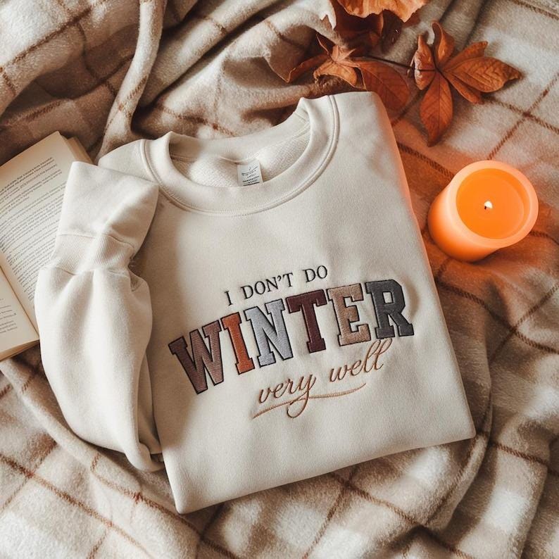 Puede incluir: Sudadera color crema con la frase bordada "I DON'T DO WINTER very well." La sudadera est&aacute; doblada sobre una manta a cuadros, con una vela naranja encendida y un libro. Hojas de oto&ntilde;o completan la escena.