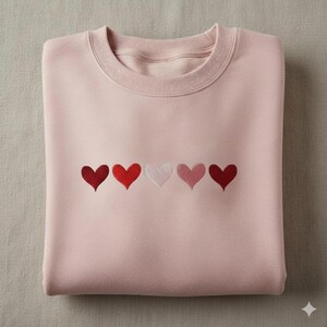 K&ouml;nnte beinhalten: Ein gefaltetes, hellrosa Sweatshirt mit Rundhalsausschnitt. Das Sweatshirt hat f&uuml;nf gestickte Herzen in Rot- und Rosat&ouml;nen auf der Brust. Die Herzen sind in einer horizontalen Linie angeordnet.