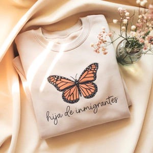 Puede incluir: Sudadera crema con un diseño de mariposa monarca en naranja y negro. La frase "hija de inmigrantes" está impresa debajo. La sudadera está doblada sobre una tela crema.