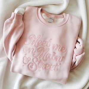 Może przedstawiać: Jasnoróżowa bluza z haftowanym napisem "Meet me on Main Street" w ozdobnej czcionce. Projekt zawiera głowę Myszki Miki i detale gwiazd. Bluza jest złożona na białym kocu.