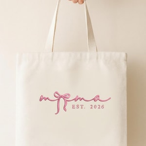 May include: Cream tote bag featuring a pink cursive "mema" design and a bow. Below the word "mema" is the text "EST. 2026" in pink. The bag is held by a hand.