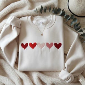 K&ouml;nnte beinhalten: Cremefarbenes Sweatshirt mit gestickten Herzen in Rot- und Rosat&ouml;nen. Der Pullover mit Rundhalsausschnitt hat lange &Auml;rmel und eine goldene Halskette. Das Sweatshirt liegt auf einer weichen, cremefarbenen Decke.