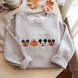 May include: A beige crewneck sweatshirt with embroidered Halloween-themed designs. The design includes two jack-o'-lanterns, a pumpkin, and a Mickey Mouse face. The sleeves are cuffed. The sweatshirt is on a light-colored surface.