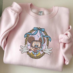 May include: A light pink sweatshirt featuring an embroidered design of Mickey Mouse wearing a snorkel and goggles. The design is framed by a scalloped border and a light blue bow. The sweatshirt has long sleeves and a crew neck.