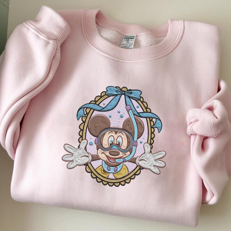 May include: A light pink sweatshirt featuring an embroidered design of Mickey Mouse wearing a snorkel and goggles. The design is framed by a scalloped border and a light blue bow. The sweatshirt has long sleeves and a crew neck.