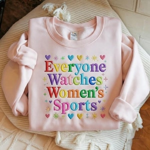 May include: A pale pink sweatshirt featuring the embroidered phrase "Everyone Watches Women's Sports" in rainbow hues. The design incorporates hearts, stars, and plus signs. The sweatshirt has long sleeves and a crew neck.