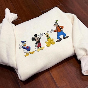 May include: Cream-colored sweatshirt featuring embroidered Disney characters: Donald Duck, Mickey Mouse, Pluto, and Goofy. The characters are in vibrant colors against the neutral background. The sweatshirt has ribbed cuffs and a crew neck.