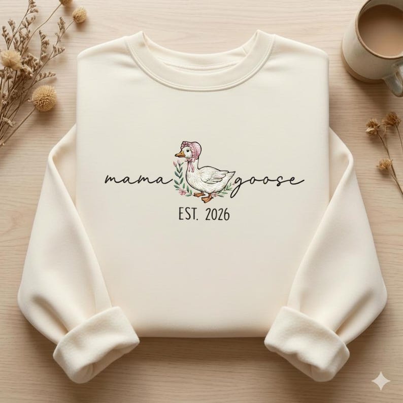 May include: Cream-colored sweatshirt with the words "mama goose" in a script font, accompanied by an illustration of a goose wearing a bonnet. Below the goose is the text "EST. 2026". The sweatshirt has a crew neck and long sleeves with rolled cuffs.