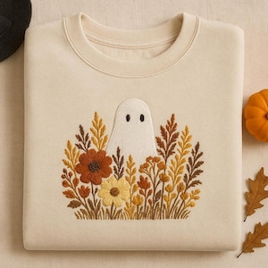 May include: Cream-colored sweatshirt with an embroidered design of a white ghost peeking out from behind a cluster of autumn-colored flowers and foliage. The design includes orange, yellow, and brown floral elements. The sweatshirt has a crew neck.