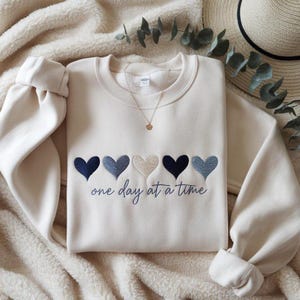 May include: Cream-colored sweatshirt with embroidered hearts in shades of blue and beige. The phrase "one day at a time" is embroidered below the hearts. A gold necklace with a small pendant is visible.