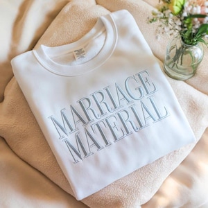 Pode incluir: Um moletom branco com as palavras "MARRIAGE MATERIAL" bordadas em azul claro. O moletom está dobrado sobre um cobertor bege, com um pequeno vaso de vidro com flores ao fundo.