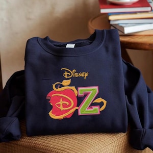 May include: A navy blue sweatshirt with an embroidered design. The design includes the word "Disney" in gold script, a red apple with a bite taken out, and the letter "Z" in green with a pink outline. The sweatshirt is on a patterned surface.