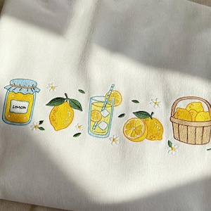 May include: White fabric featuring an embroidered lemon design. The design includes a jar of lemon preserves, a whole lemon, a glass of lemonade with ice, and a basket of lemons. Small white flowers and green leaves are also embroidered.