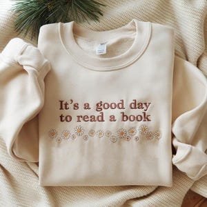 Op de afbeelding: Crèmekleurige sweatshirt met de geborduurde tekst "It's a good day to read a book" in bruine letters. Een rij geborduurde madeliefjes in wit en geel accentueert de tekst. De sweatshirt is opgevouwen op een crèmekleurige deken.