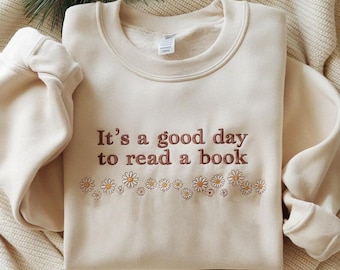 May include: Cream-colored sweatshirt with the embroidered phrase "It's a good day to read a book" in brown lettering. A row of embroidered daisies in white and yellow accents the text. The sweatshirt is folded on a cream-colored blanket.